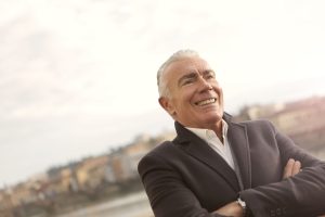 A senior man in business attire smiles confidently during a sunny outdoor setting.